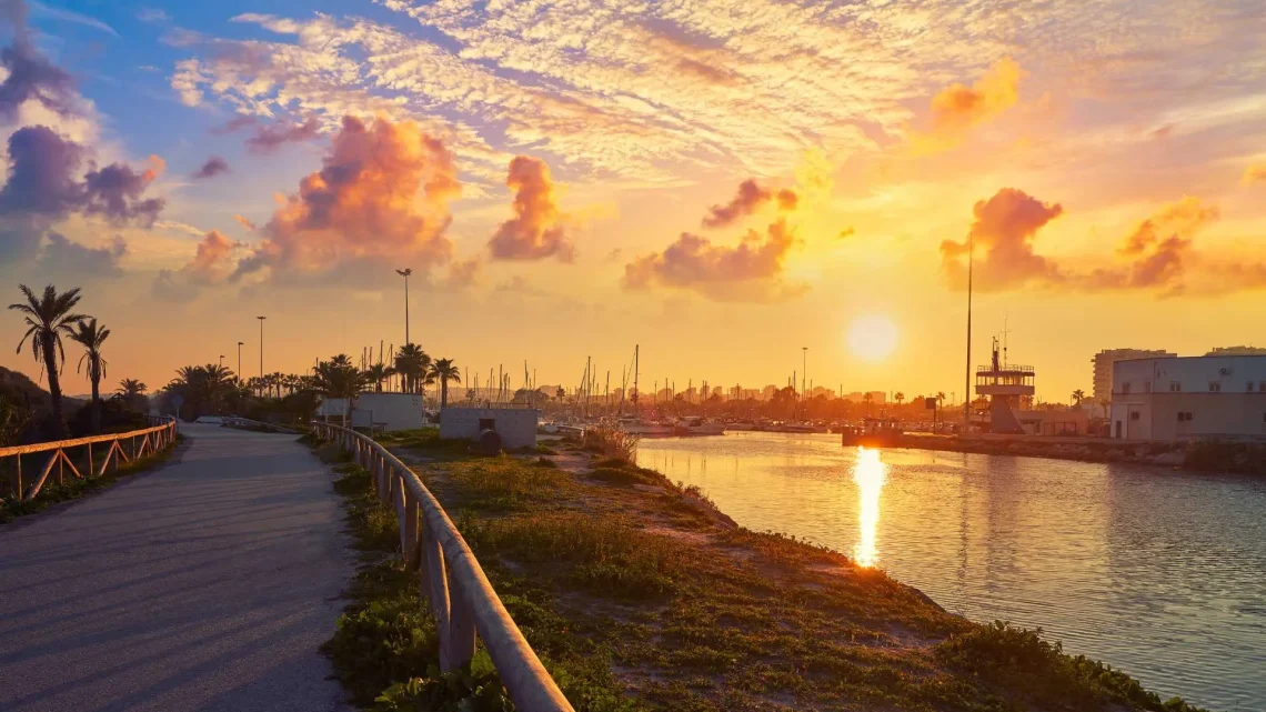 Promenade entlang eines Hafens bei Sonnenuntergang mit Wasser, Booten und Palmen am Meer.